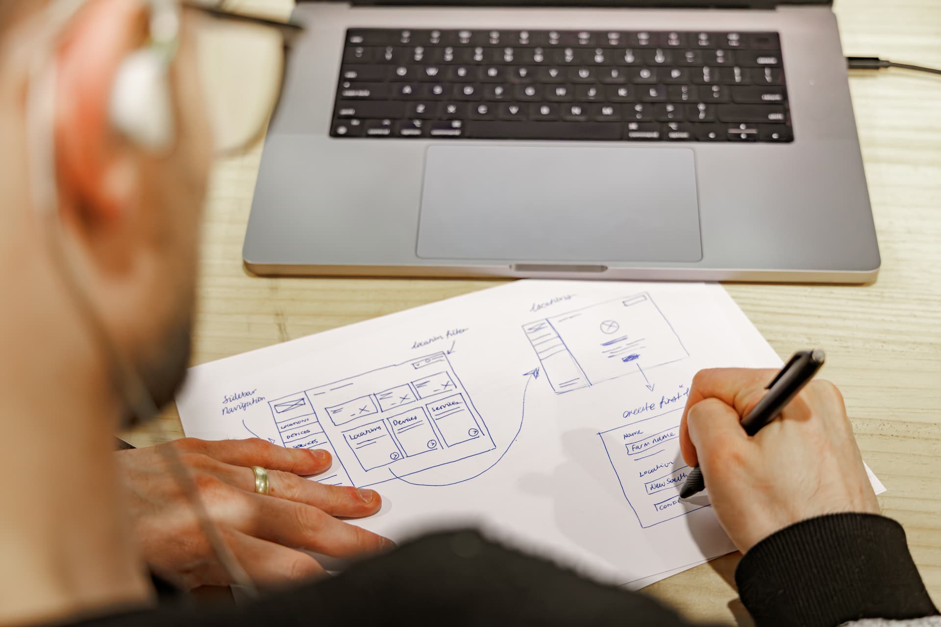 A designer sketching interface wireframes on paper beside a laptop