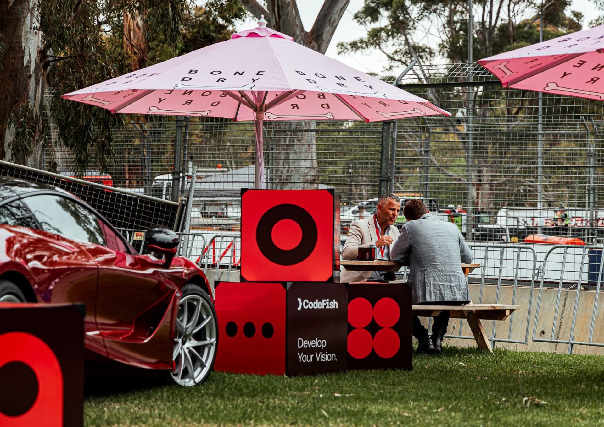 Two men talking at a picnic table under a pink umbrella, beside CodeFish Develop Your Vision cubes and a red sports car