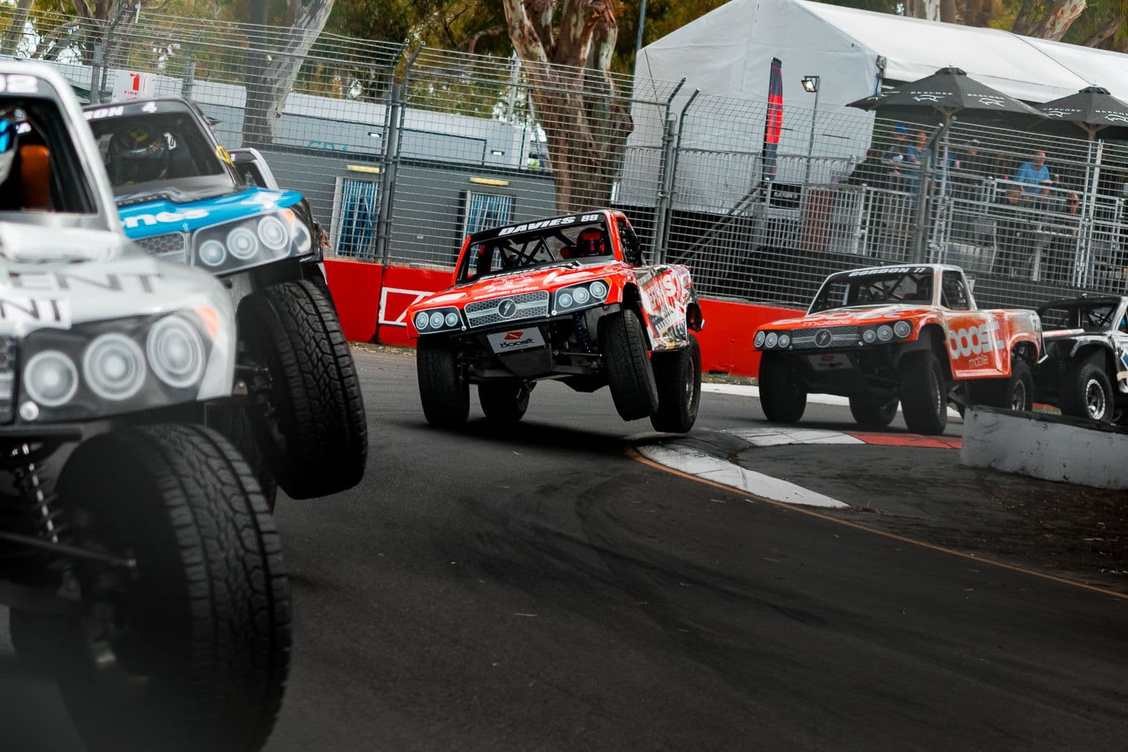 Pack of stadium super trucks racing through a tree-lined corner of a street circuit